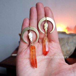 Könnte beinhalten: Ein Paar goldene Halbmond-Ohrringe mit einem kleinen Feder-Anhänger und einer baumelnden orangefarbenen Kristallspitze.