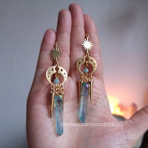 May include: Gold earrings with a sun and crescent moon design, featuring a blue crystal pendant and a small blue bead. The earrings are hanging from a hand.