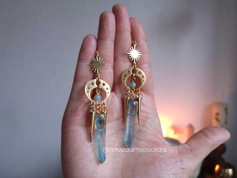May include: Gold earrings with a sun and crescent moon design, featuring a blue crystal pendant and a small blue bead. The earrings are hanging from a hand.