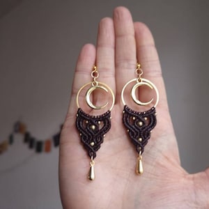 May include: A pair of brown and gold earrings with a crescent moon design. The earrings are made with macrame and have a gold-toned metal hoop and a teardrop-shaped pendant.