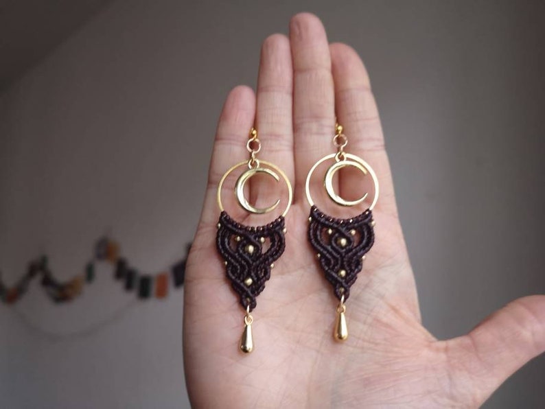 May include: A pair of brown and gold earrings with a crescent moon design. The earrings are made with macrame and have a gold-toned metal hoop and a teardrop-shaped pendant.