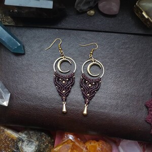 May include: A pair of maroon and gold macrame earrings with a crescent moon charm. The earrings are hanging from a dark brown leather surface.