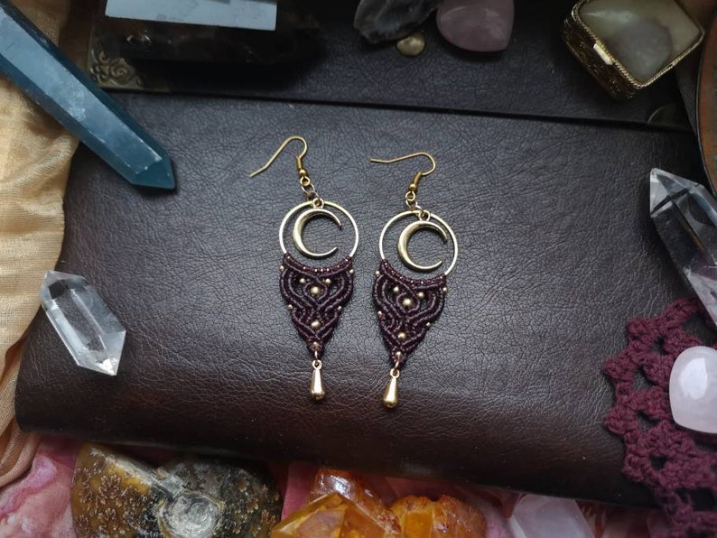 May include: A pair of maroon and gold macrame earrings with a crescent moon charm. The earrings are hanging from a dark brown leather surface.
