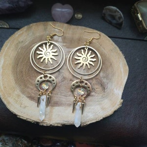 May include: Gold earrings with a sun and moon design. The earrings feature a circle with a sun design and a crescent moon with a crystal charm. The earrings are hanging from a gold hook.