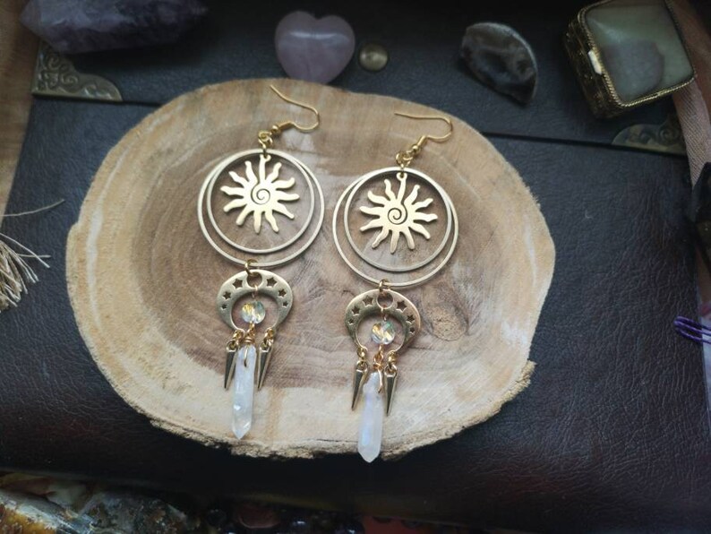 May include: Gold earrings with a sun and moon design. The earrings feature a circle with a sun design and a crescent moon with a crystal charm. The earrings are hanging from a gold hook.