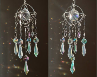 Puede incluir: Un atrapasol de cristal plateado con un prisma de cristal redondo enjaulado y múltiples gotas de cristal iridiscente que cuelgan.