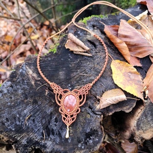 May include: A brown macrame necklace with a red gemstone pendant. The necklace is on a wooden surface with autumn leaves.
