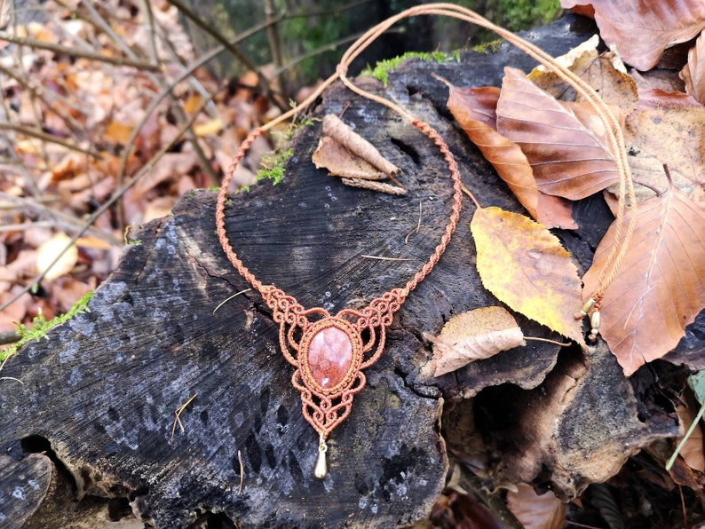 Puede incluir: Un collar de macram&eacute; marr&oacute;n con un colgante de piedra roja. El collar est&aacute; sobre una superficie de madera con hojas de oto&ntilde;o.