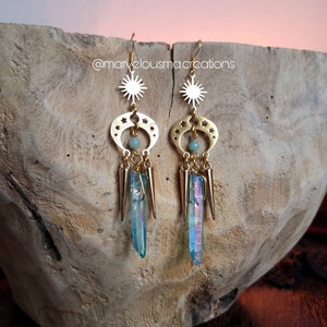 May include: Gold earrings with a crescent moon and star design, featuring iridescent blue crystal accents and a green stone. The earrings are displayed on a wooden surface. @marvelousmacreations