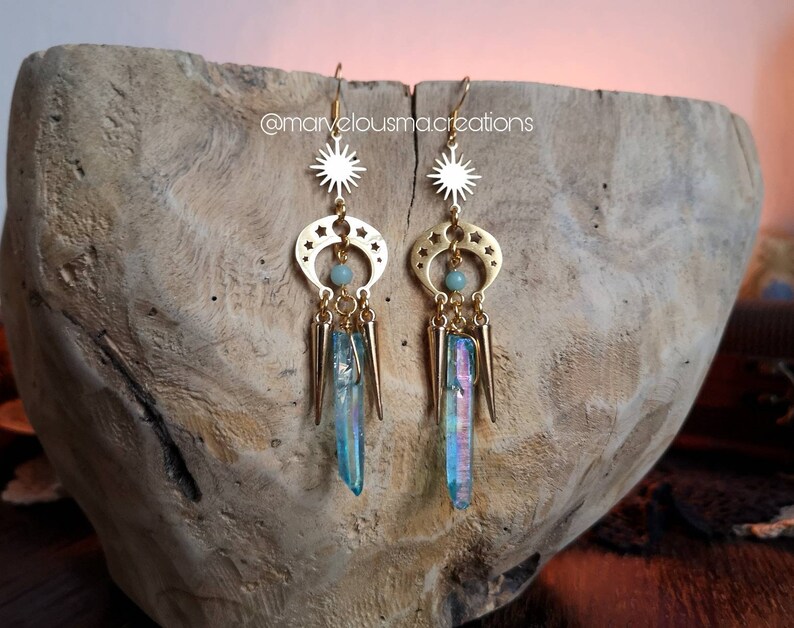 May include: Gold earrings with a crescent moon and star design, featuring iridescent blue crystal accents and a green stone. The earrings are displayed on a wooden surface. @marvelousmacreations