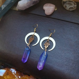 Puede incluir: Un par de pendientes de oro con un dise&ntilde;o de media luna y un amuleto de mano de hamsa. Cada pendiente presenta un colgante de cristal morado y azul.