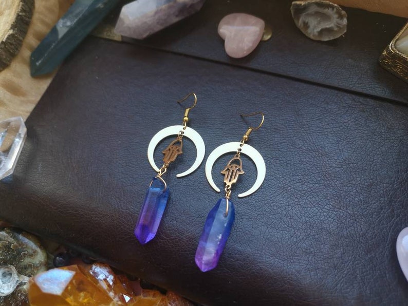 Puede incluir: Un par de pendientes de oro con un dise&ntilde;o de media luna y un amuleto de mano de hamsa. Cada pendiente presenta un colgante de cristal morado y azul.