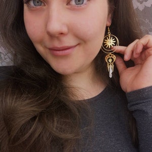 May include: Gold tone earrings with a sun and moon design. The sun is in the center of a circle and the moon is hanging from the bottom of the circle. There are small white beads hanging from the moon.