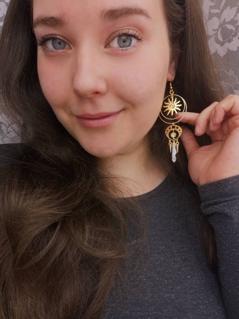 May include: Gold tone earrings with a sun and moon design. The sun is in the center of a circle and the moon is hanging from the bottom of the circle. There are small white beads hanging from the moon.