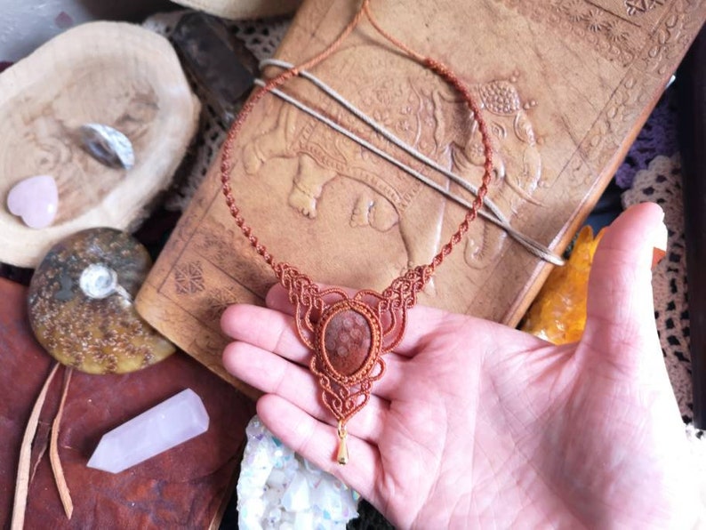 Puede incluir: Un collar de cord&oacute;n de cuero marr&oacute;n con un dise&ntilde;o anudado y un gran colgante redondo de piedra marr&oacute;n.
