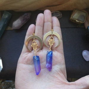 Puede incluir: Pendientes de media luna dorados con un amuleto de mano de hamsa y un colgante de cristal azul y morado.
