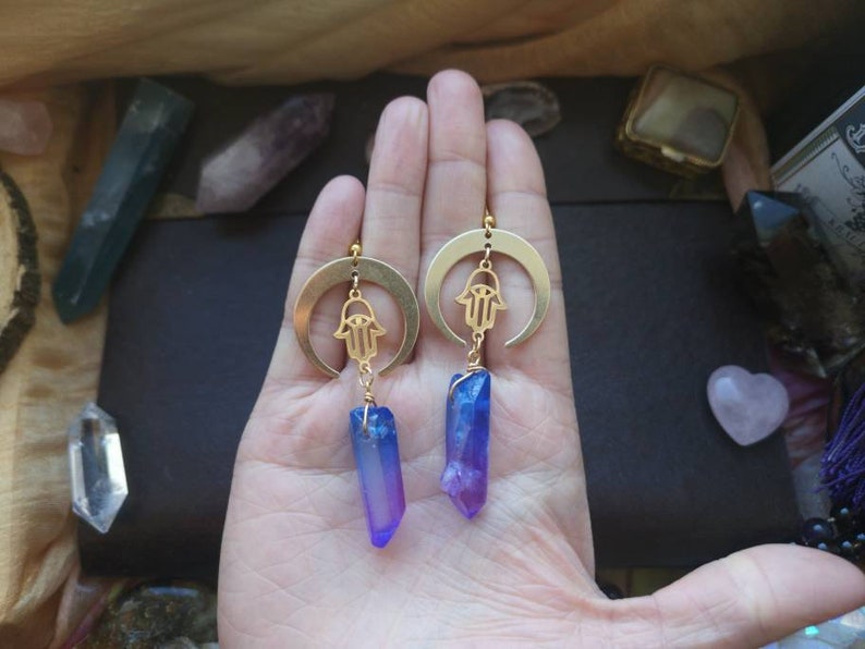 Puede incluir: Pendientes de media luna dorados con un amuleto de mano de hamsa y un colgante de cristal azul y morado.
