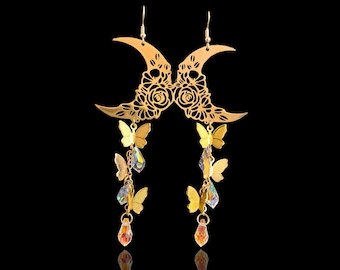May include: Gold-toned earrings featuring crescent moon shapes with floral cutouts. Dangling from the moons are gold butterflies and iridescent crystal accents, culminating in teardrop-shaped orange crystals. The earrings hang against a black background.
