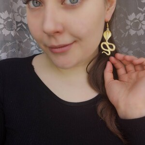 May include: Gold snake earrings with a detailed design. The earrings are dangling and have a hook closure.