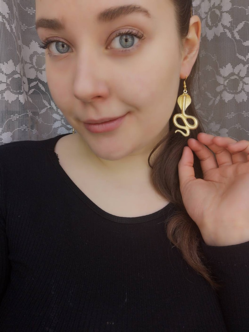 May include: Gold snake earrings with a detailed design. The earrings are dangling and have a hook closure.