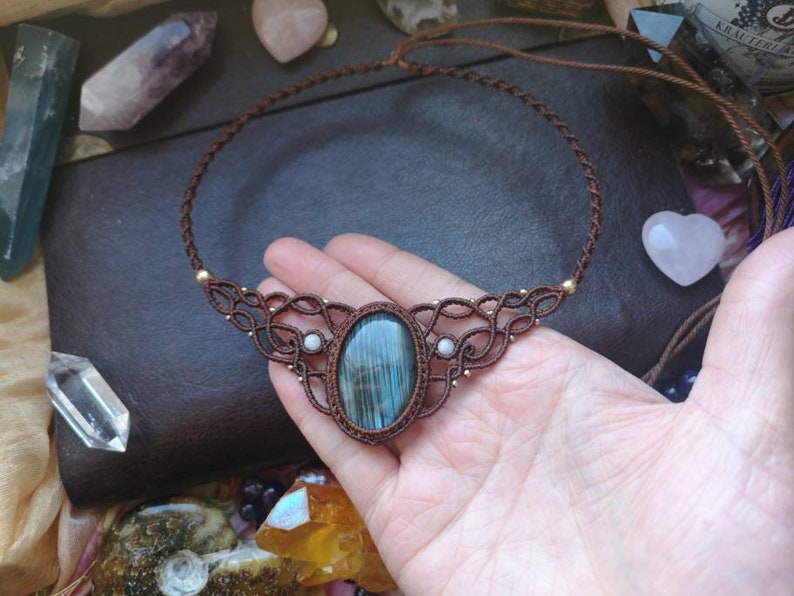 May include: A brown cord necklace with a large oval labradorite gemstone pendant and two smaller white gemstone accents. The necklace is knotted with a macrame design.
