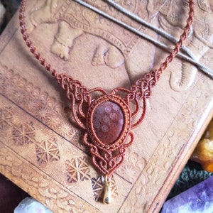 Puede incluir: Un collar de macram&eacute; marr&oacute;n con un colgante de piedra roja y marr&oacute;n. El collar est&aacute; sobre un fondo de cuero marr&oacute;n.