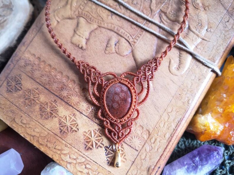 Puede incluir: Un collar de macram&eacute; marr&oacute;n con un colgante de piedra roja y marr&oacute;n. El collar est&aacute; sobre un fondo de cuero marr&oacute;n.