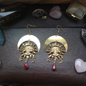 May include: Gold tone earrings featuring a crescent moon and sun face with red gemstone accents.