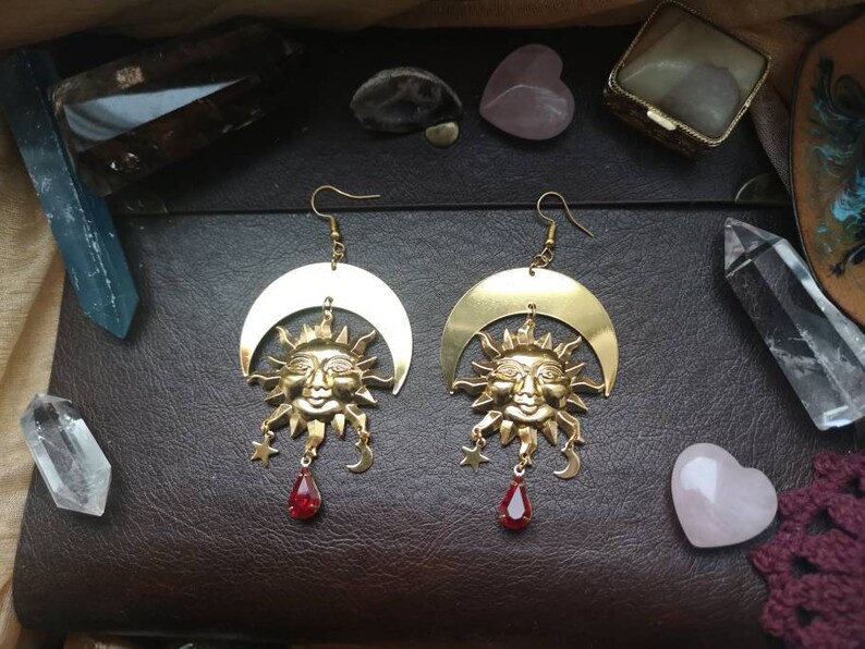 May include: Gold tone earrings featuring a crescent moon and sun face with red gemstone accents.