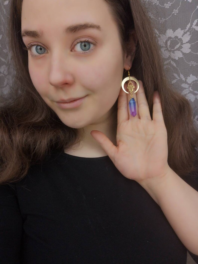 Puede incluir: Pendientes de oro con un creciente de luna y un amuleto de mano de hamsa, colgando de un cristal morado y azul.