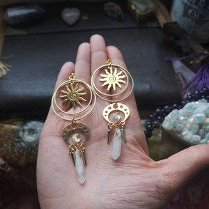 May include: Gold earrings with a sun and moon design. Each earring features a circular sun with a spiral design, a crescent moon with stars, and a dangling crystal.