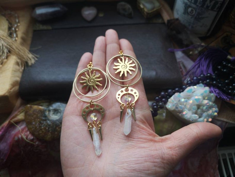 May include: Gold earrings with a sun and moon design. Each earring features a circular sun with a spiral design, a crescent moon with stars, and a dangling crystal.