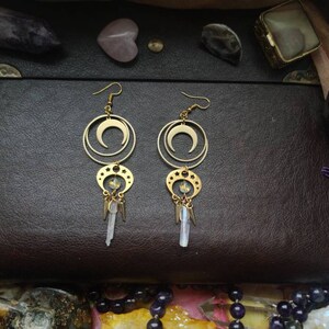 May include: Gold crescent moon earrings with dangling crystal charms. The earrings are on a brown leather background.