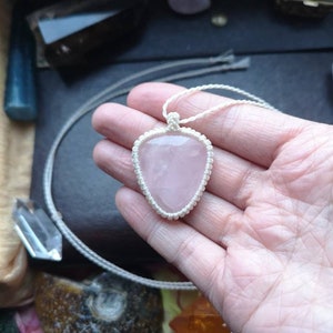 May include: A pink rose quartz heart pendant on a white cord necklace. The pendant is held in a hand.