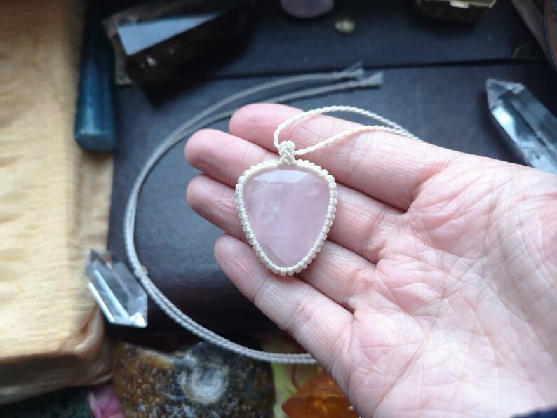 May include: A pink rose quartz heart pendant on a white cord necklace. The pendant is held in a hand.