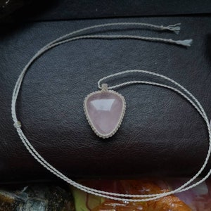 May include: A pink rose quartz pendant necklace with a white cord. The pendant is in the shape of a heart and is surrounded by a white cord.