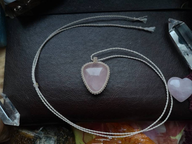 May include: A pink rose quartz pendant necklace with a white cord. The pendant is in the shape of a heart and is surrounded by a white cord.