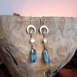 K&ouml;nnte beinhalten: Goldene Ohrringe mit einem Halbmond, einem Stern und einem blauen Kristall-Anh&auml;nger. Die Ohrringe h&auml;ngen an einer Holzschale.