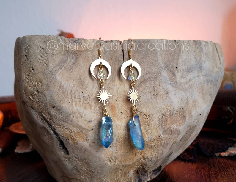 K&ouml;nnte beinhalten: Goldene Ohrringe mit einem Halbmond, einem Stern und einem blauen Kristall-Anh&auml;nger. Die Ohrringe h&auml;ngen an einer Holzschale.