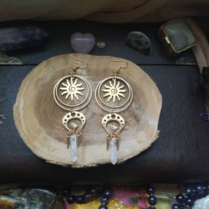 May include: Gold tone earrings with a sun and moon design. The earrings feature a circular pendant with a sun design and a crescent moon pendant with a crystal charm. The earrings are hanging from a gold tone chain.