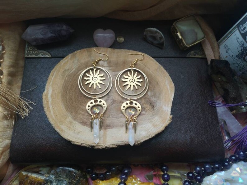 May include: Gold tone earrings with a sun and moon design. The earrings feature a circular pendant with a sun design and a crescent moon pendant with a crystal charm. The earrings are hanging from a gold tone chain.