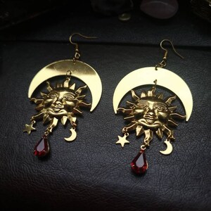 May include: Gold earrings with a crescent moon and sun face design. Each earring features a red gemstone dangle.