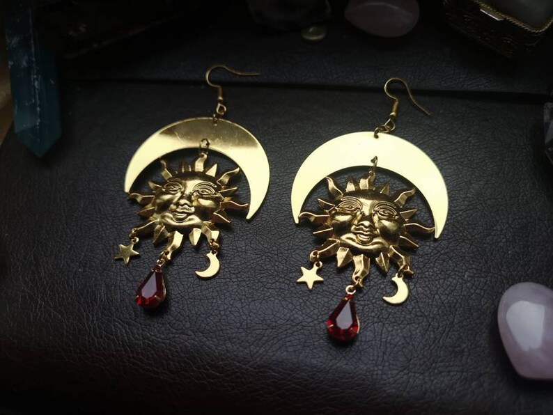 May include: Gold earrings with a crescent moon and sun face design. Each earring features a red gemstone dangle.