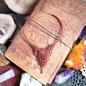Puede incluir: Un collar con un colgante de piedra rojo-marr&oacute;n y un dise&ntilde;o de cord&oacute;n anudado. El collar se muestra en un diario encuadernado en cuero con intrincados patrones en relieve.