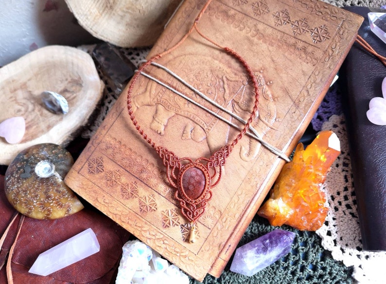 Puede incluir: Un collar con un colgante de piedra rojo-marr&oacute;n y un dise&ntilde;o de cord&oacute;n anudado. El collar se muestra en un diario encuadernado en cuero con intrincados patrones en relieve.