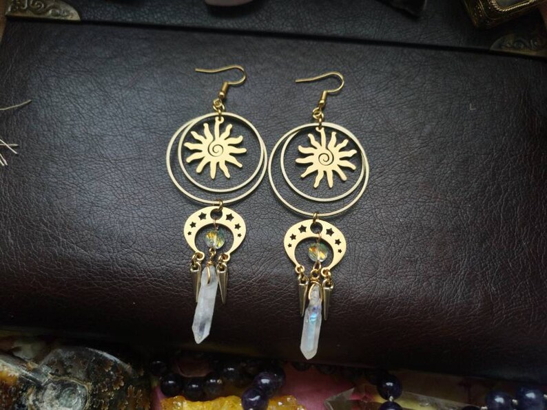 May include: Gold earrings with a sun and moon design. The earrings feature a circle with a sun design and a crescent moon with stars. The earrings are accented with clear crystals.