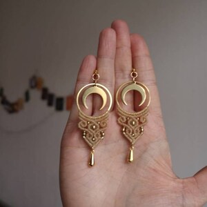 May include: A pair of gold earrings with a crescent moon charm and a delicate macrame design. The earrings are hanging from a hand.