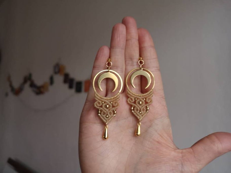 May include: A pair of gold earrings with a crescent moon charm and a delicate macrame design. The earrings are hanging from a hand.