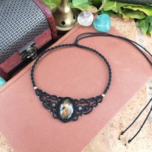 以下が含まれることがあります： 大きな楕円形のラブラドライトの宝石のペンダントが付いた黒いマクラメチョーカーネックレス。ネックレスは金色のビーズが付いており、黒い紐で結ばれています。