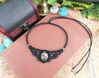 Puede incluir: Un collar gargantilla de macramé negro con un colgante de labradorita ovalado grande. El collar tiene cuentas doradas y está atado con un cordón negro.